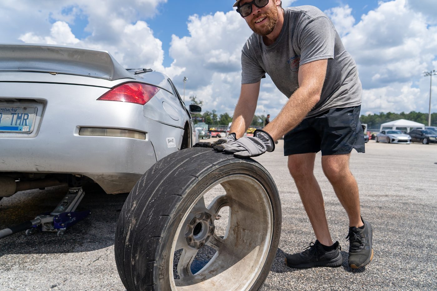 Crew member rolling a worn drift tire past a jacked-up car in the pits